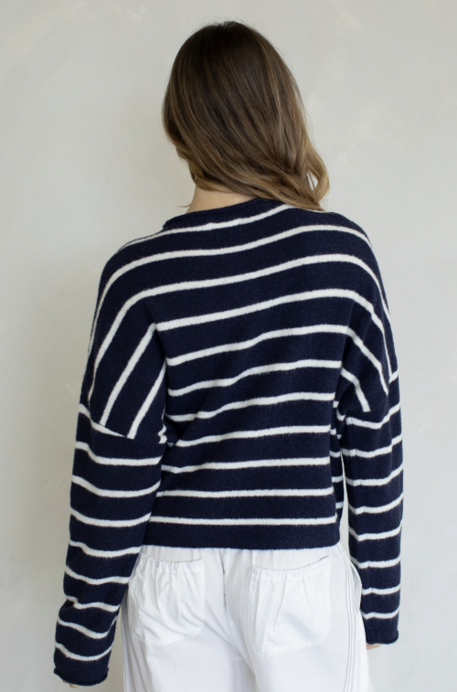 Woman shown from behind wearing navy and white stripe cardigan and white pants on plain background