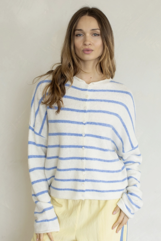 A woman wearing a cream knit cardigan with thin blue horizontal stripes and pale yellow pants, standing against a plain light beige wall.