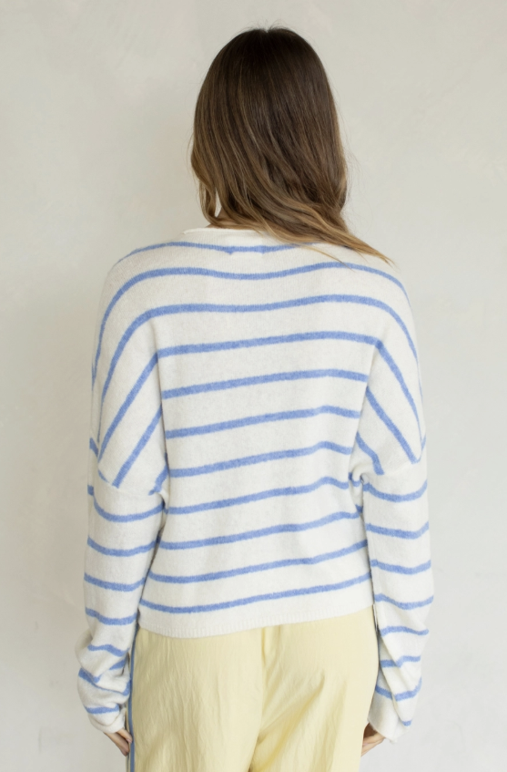 A woman viewed from behind wearing a cream knit sweater with thin blue horizontal stripes and pale yellow pants, standing against a plain light beige wall.