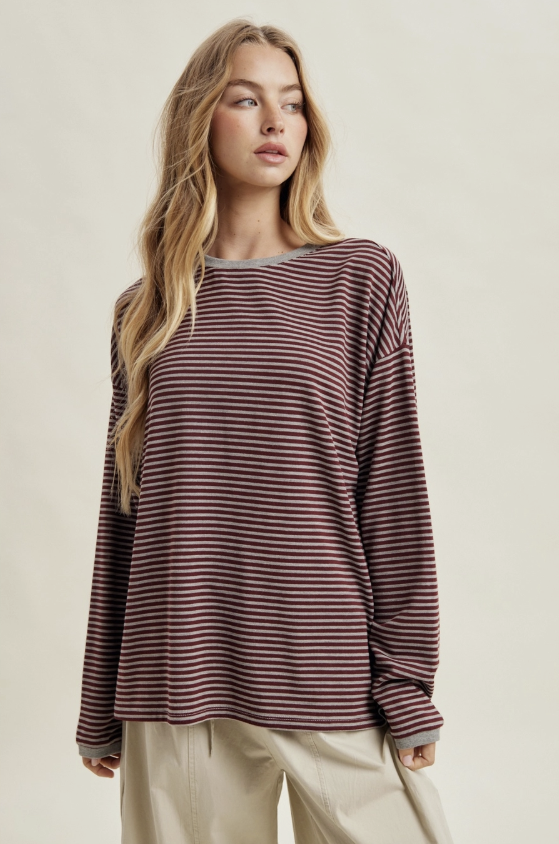 A woman modeling a long-sleeve crewneck top with thin horizontal burgundy and dark brown stripes, paired with light beige pants, against a solid light beige background.