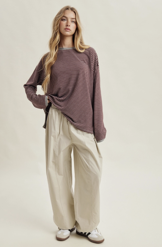A woman modeling a loose, long-sleeve striped knit top in dark red and beige, paired with wide-leg beige trousers and white sneakers, against a solid light beige studio background.