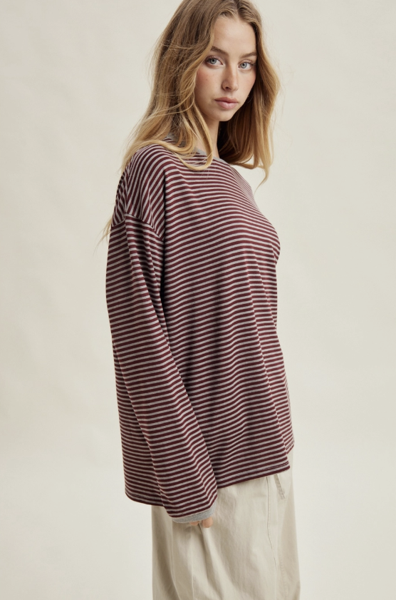 A woman wearing a long-sleeve crewneck top with thin horizontal burgundy and beige stripes, paired with light beige trousers, photographed against a plain light beige studio background.