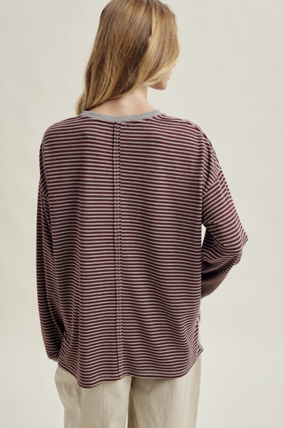 A woman viewed from the back wearing a long-sleeve top with thin horizontal dark red and brown stripes, paired with light beige pants, against a plain light beige studio background.