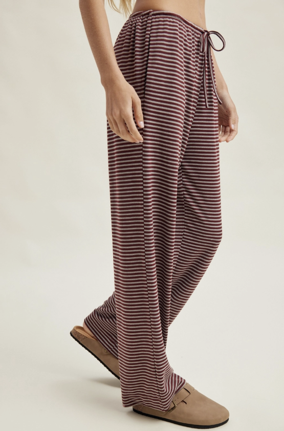 A person wearing loose, wide-leg striped lounge pants in dark burgundy and beige, paired with tan slip-on mules, photographed against a plain light beige studio background.