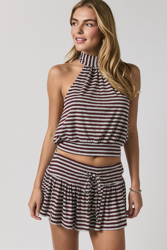 A woman modeling a matching two-piece set featuring a halter crop top and a ruffled mini skirt, both with horizontal dark red and beige stripes, against a light gray studio background.