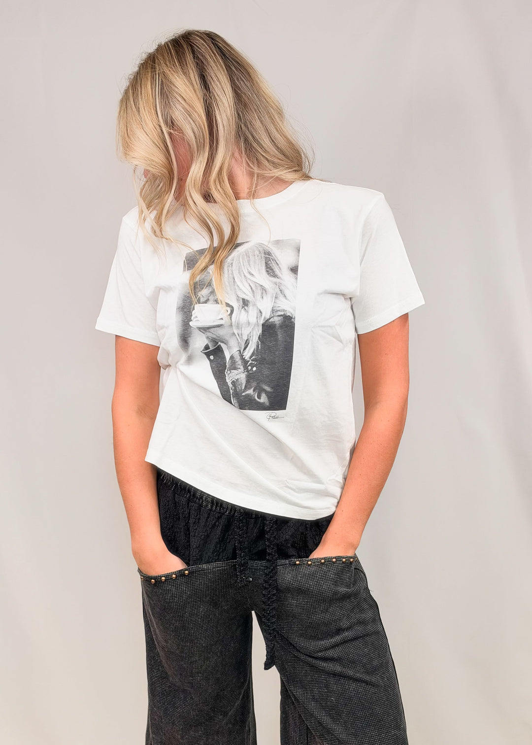 Woman wearing graphic tee of woman drinking coffee with black pants on plain background