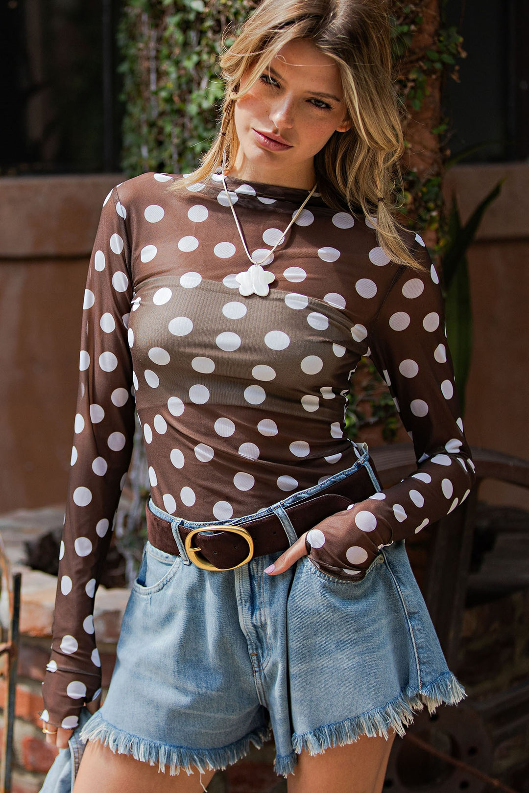 A woman wearing a sheer brown long-sleeve top with large white polka dots, paired with light blue denim shorts and a brown belt with a gold buckle.