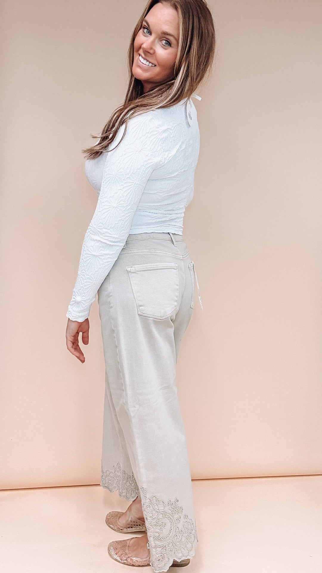 A woman modeling a light blue long-sleeve textured top and light beige wide-leg jeans with lace detailing at the hems, standing against a solid beige studio backdrop.