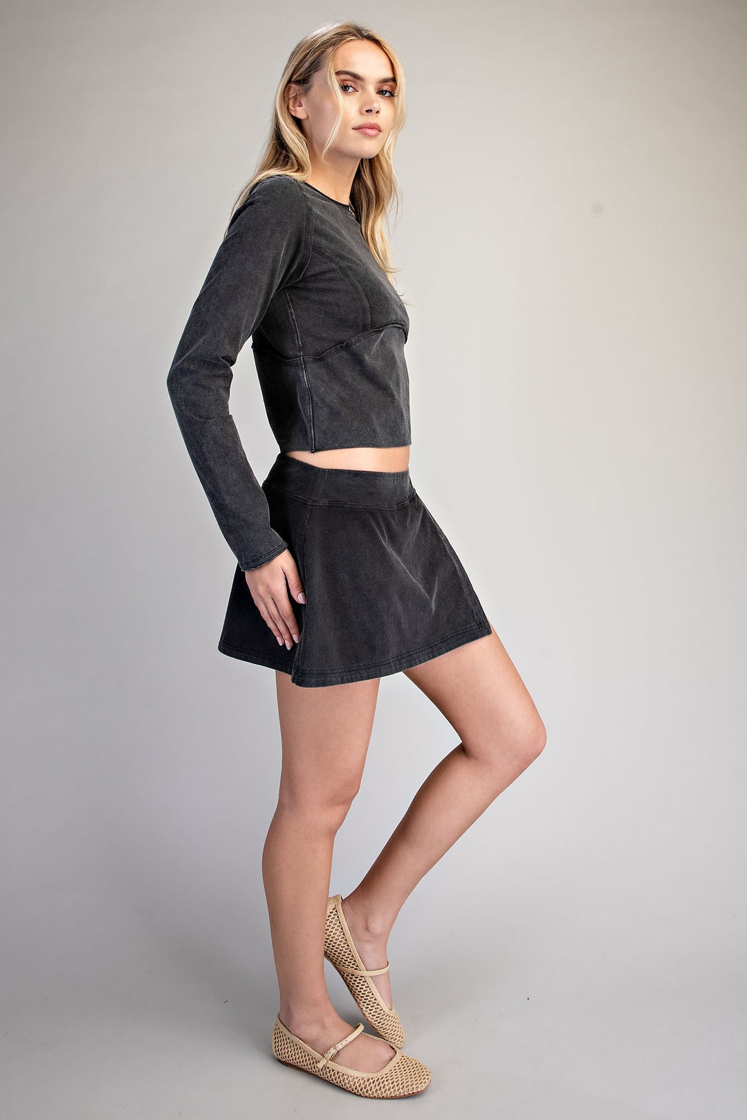 A woman modeling a dark gray corduroy two-piece set consisting of a long-sleeve crop top and a matching mini skirt, paired with beige woven flat shoes, against a neutral gray studio background.