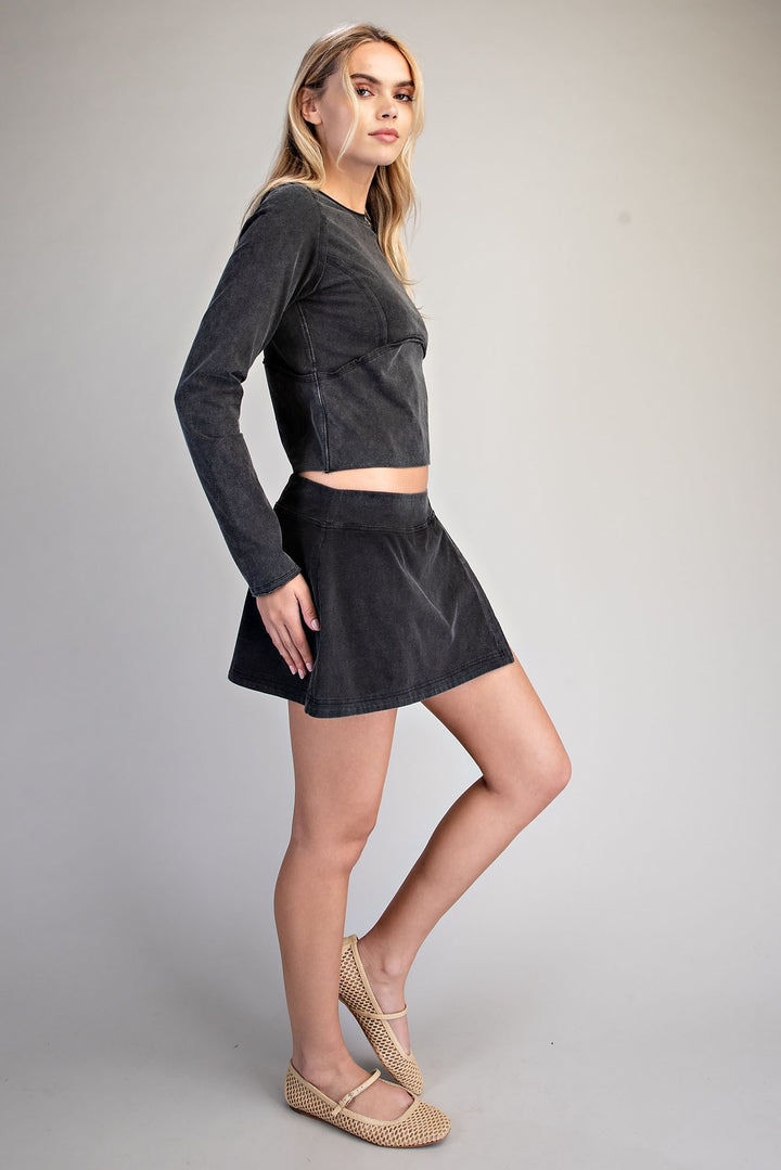 A woman modeling a dark gray corduroy two-piece set consisting of a long-sleeve crop top and a matching mini skirt, paired with beige woven flat shoes, against a neutral gray studio background.