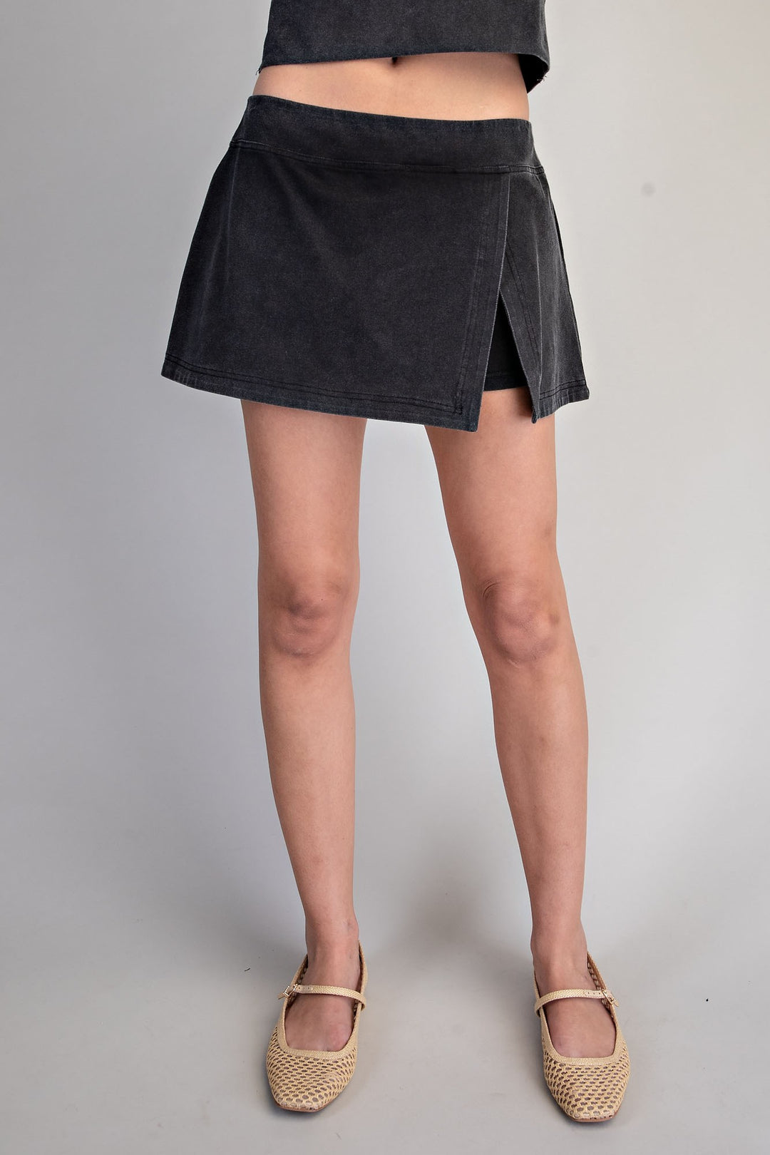 A person wearing a dark charcoal gray wrap-style mini skirt with side slits, paired with beige woven Mary Jane flats, shown from the waist down against a light gray studio background.