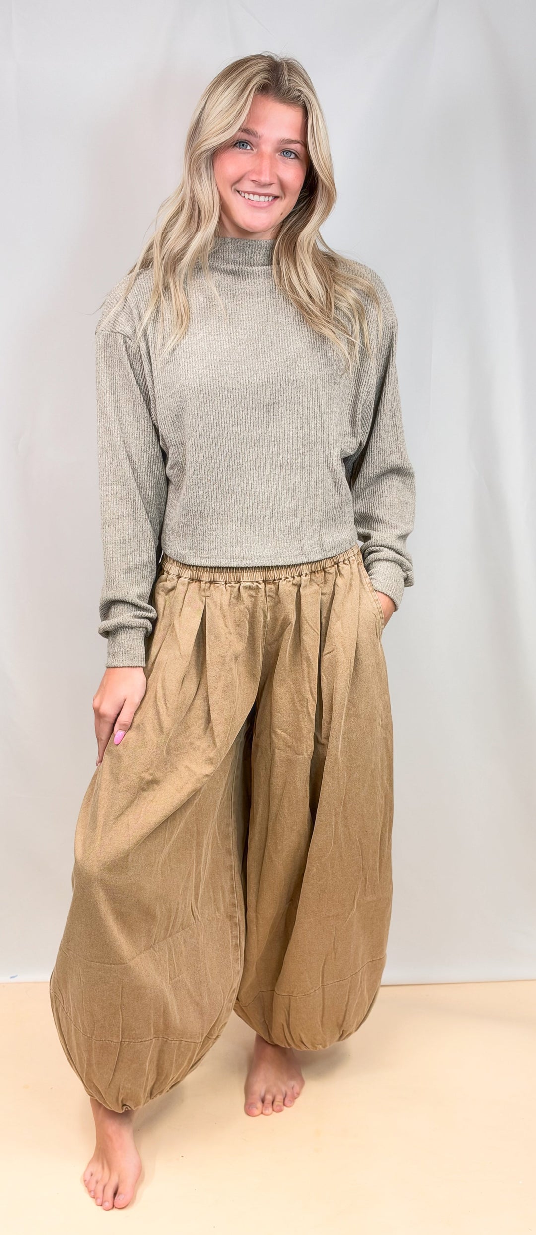 A woman stands barefoot against a light neutral studio backdrop, wearing a light gray turtleneck sweater and loose, baggy tan pants with gathered hems and side pockets.