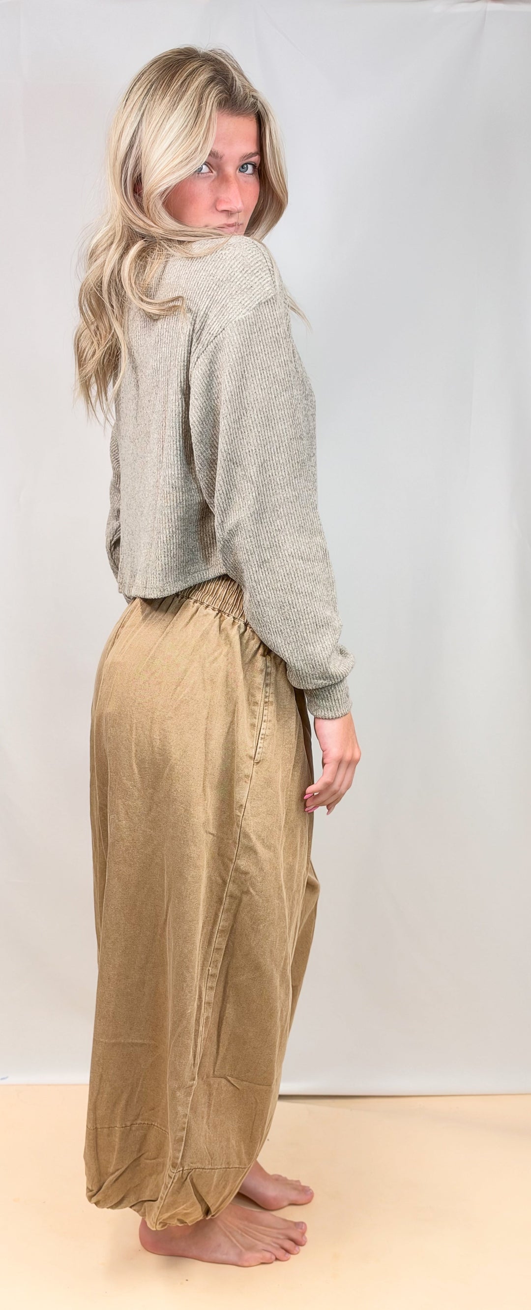 A woman stands barefoot in a studio wearing a light gray cropped sweater and loose tan pants, photographed against a plain light backdrop.