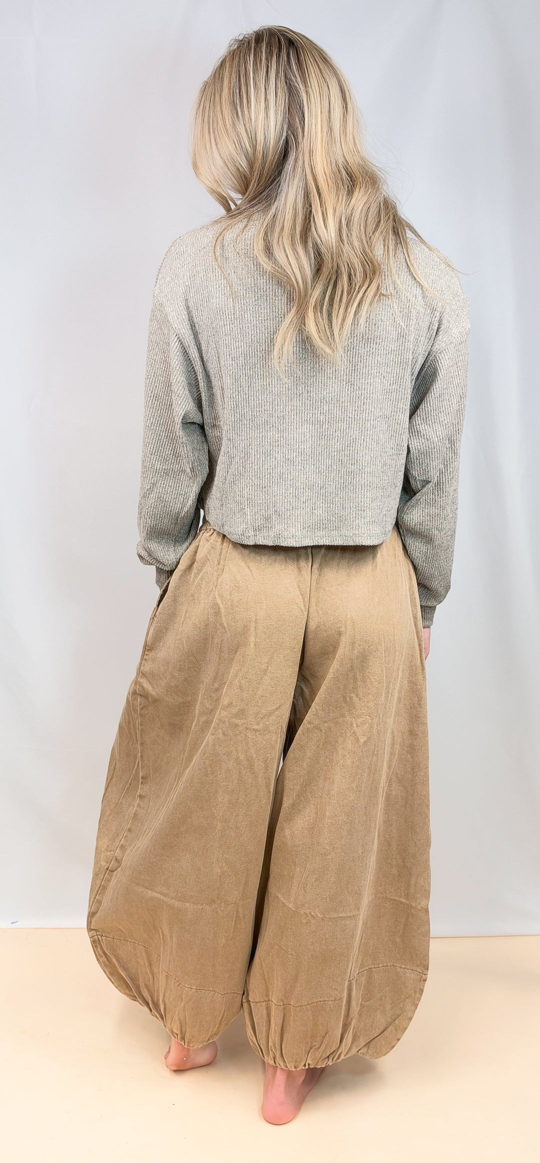 A woman shown from behind wearing a light gray long-sleeve cropped sweater and voluminous tan corduroy harem pants, standing barefoot against a plain light gray studio backdrop.