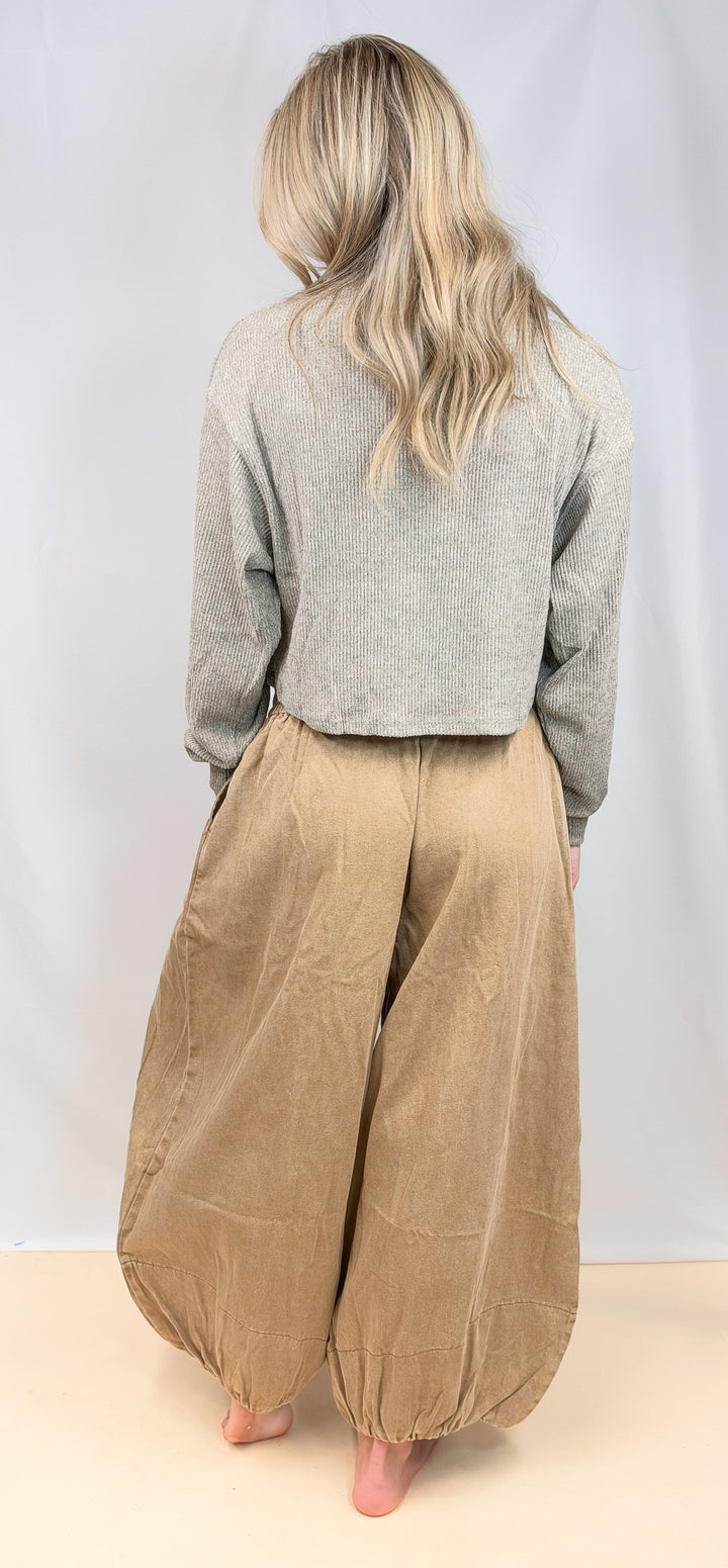 A woman shown from behind wearing a light gray long-sleeve cropped sweater and voluminous tan corduroy harem pants, standing barefoot against a plain light gray studio backdrop.