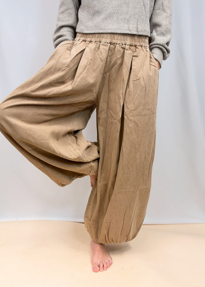 A person wearing loose, wide-leg tan pants with an elastic waistband and gathered cuffs, paired with a gray top, standing barefoot against a plain light background.