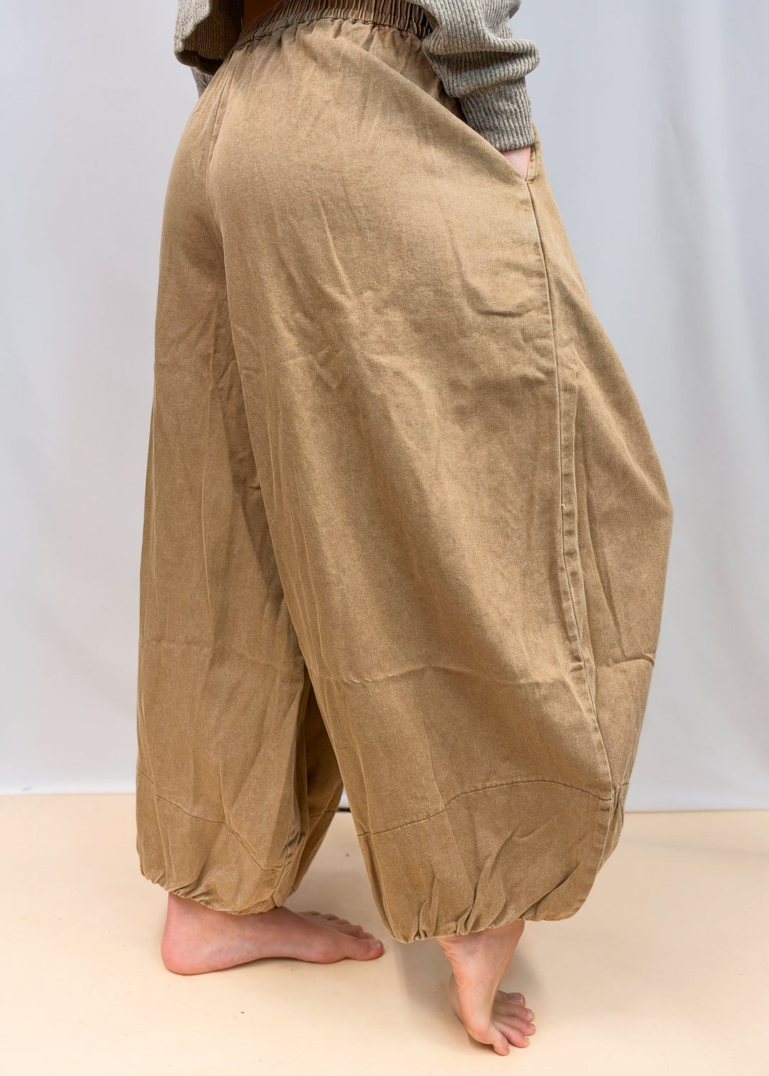 A person wearing loose, baggy tan pants with an elastic waistband and gathered cuffs, shown from the waist down against a plain light backdrop.