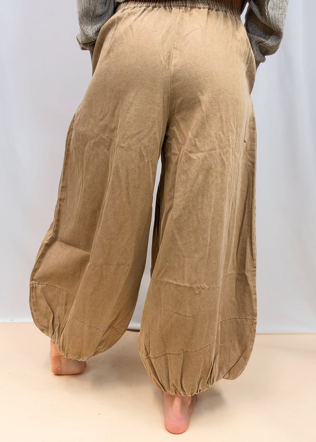 A person shown from the back wearing loose, wide-leg tan pants with gathered cuffs and a gray top, standing barefoot against a plain light background.