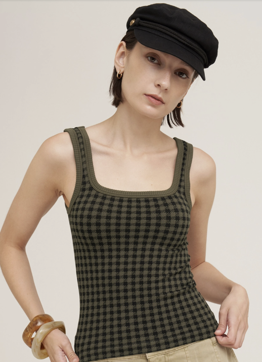 A woman wearing a black and olive green checkered knit tank top, a black newsboy cap, and beige pants, photographed against a plain light beige studio background.
