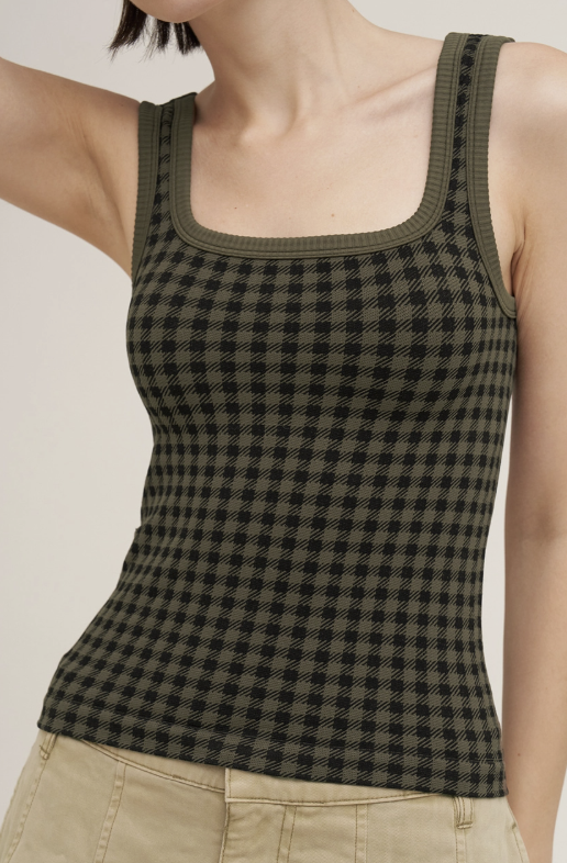 A close-up of a model wearing a ribbed knit tank top with a black and olive green checkered pattern, paired with beige pants, against a plain light background.