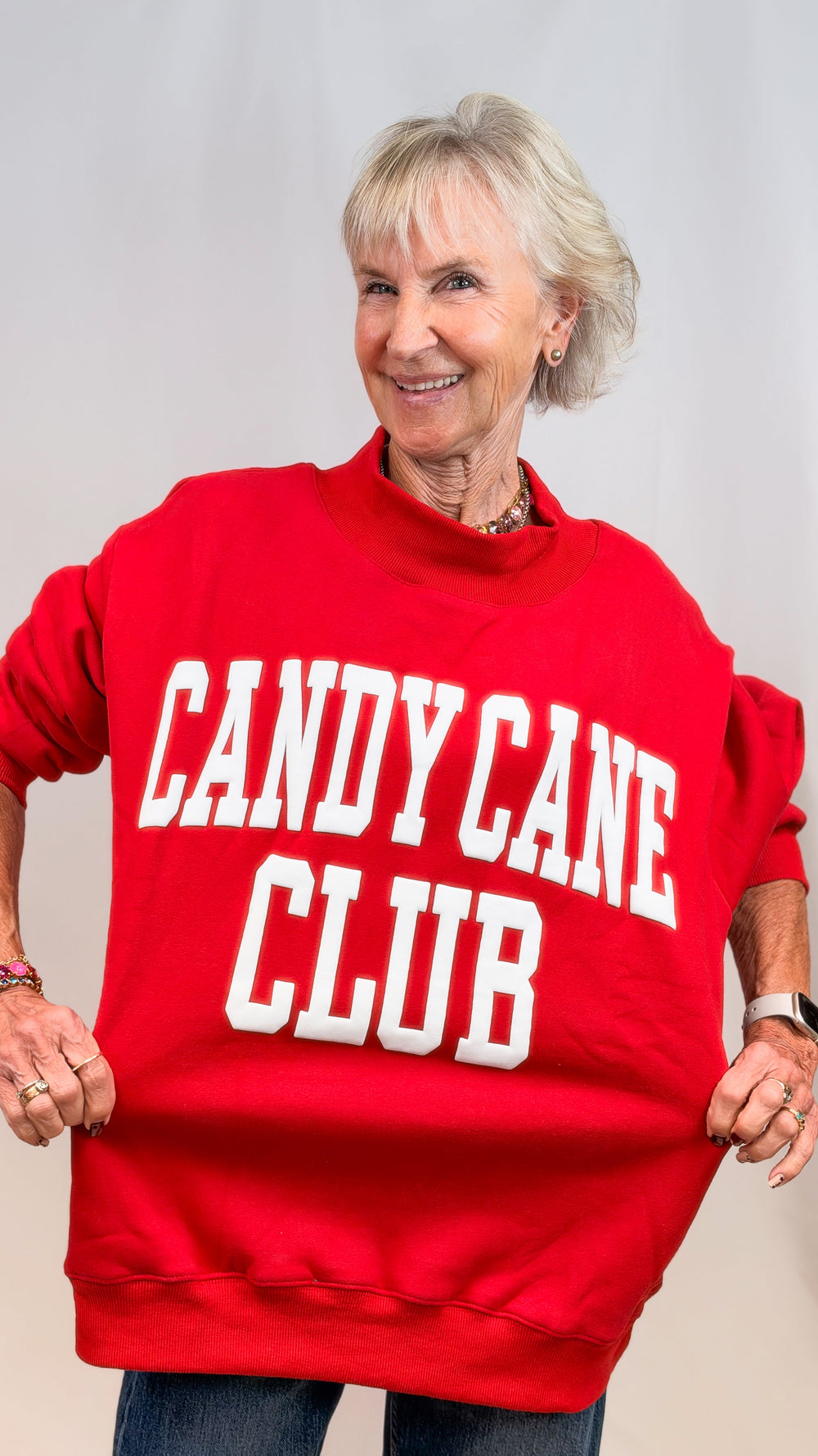 Woman wearing red mock neck sweatshirt with 'CANDY CANE CLUB' on front and jeans on plain background