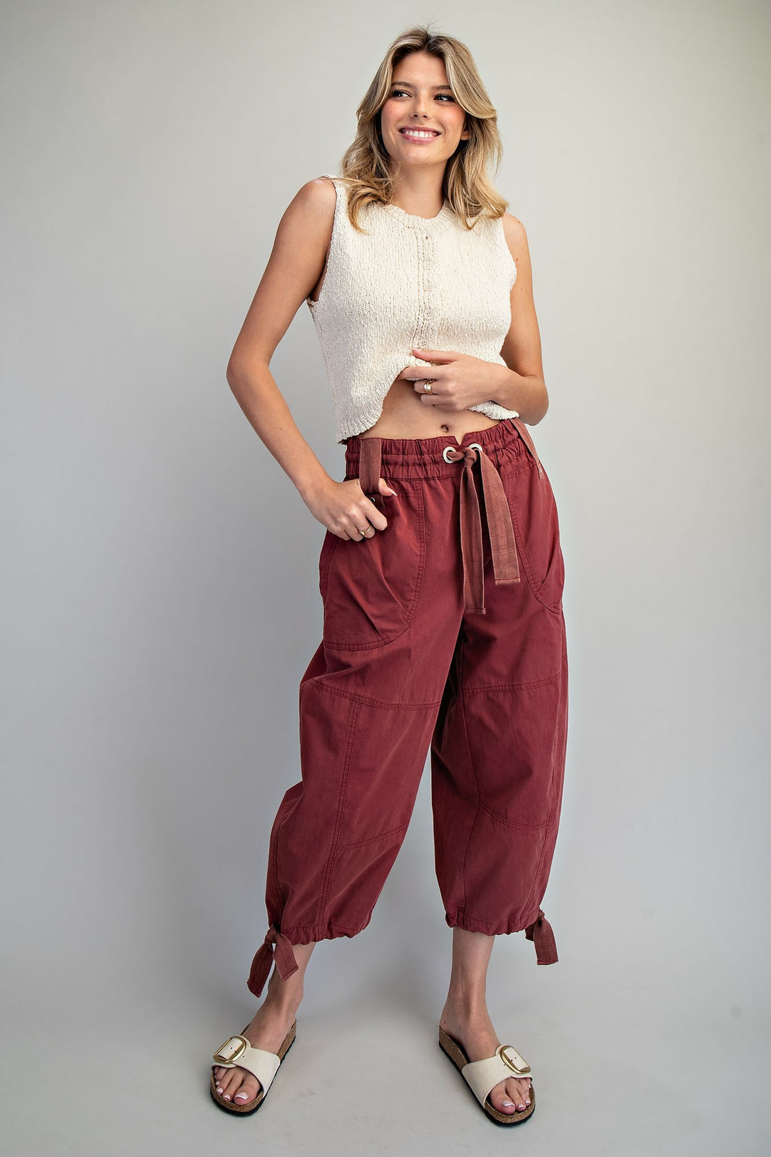 A woman modeling a pair of loose-fitting, high-waisted maroon pants with a drawstring waist, paired with a cream sleeveless top and beige slide sandals, against a solid light gray background.