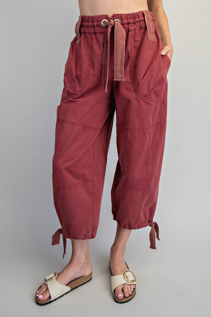A person wearing loose, cropped dusty rose pants with an elastic drawstring waist, side pockets, and tie details at the ankles, paired with cream slide sandals against a light gray studio background.