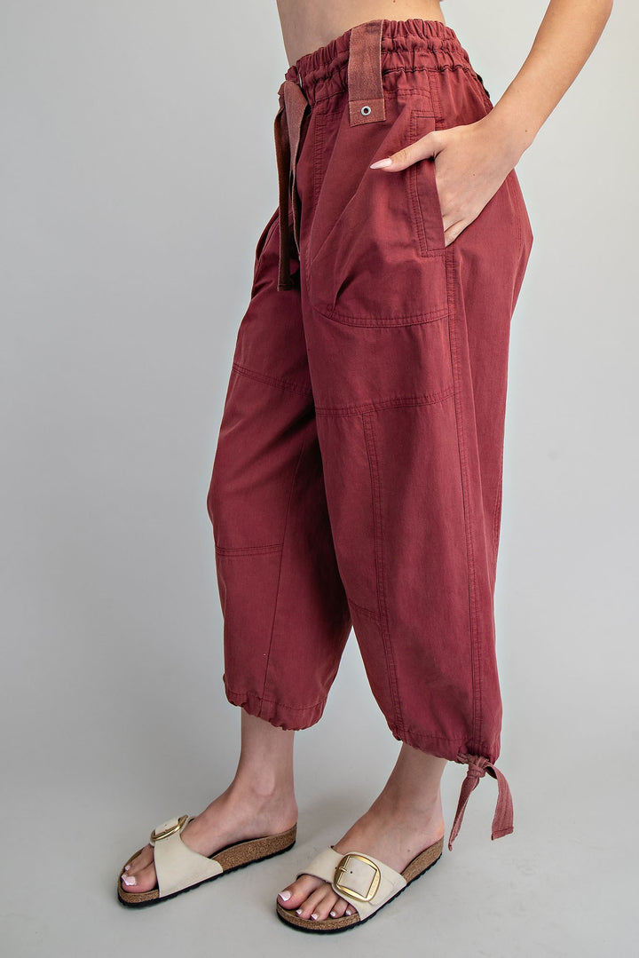 A person wearing loose, wide-leg cropped pants in a muted burgundy or dusty rose tone, paired with cream slide sandals, photographed against a plain light gray studio background.