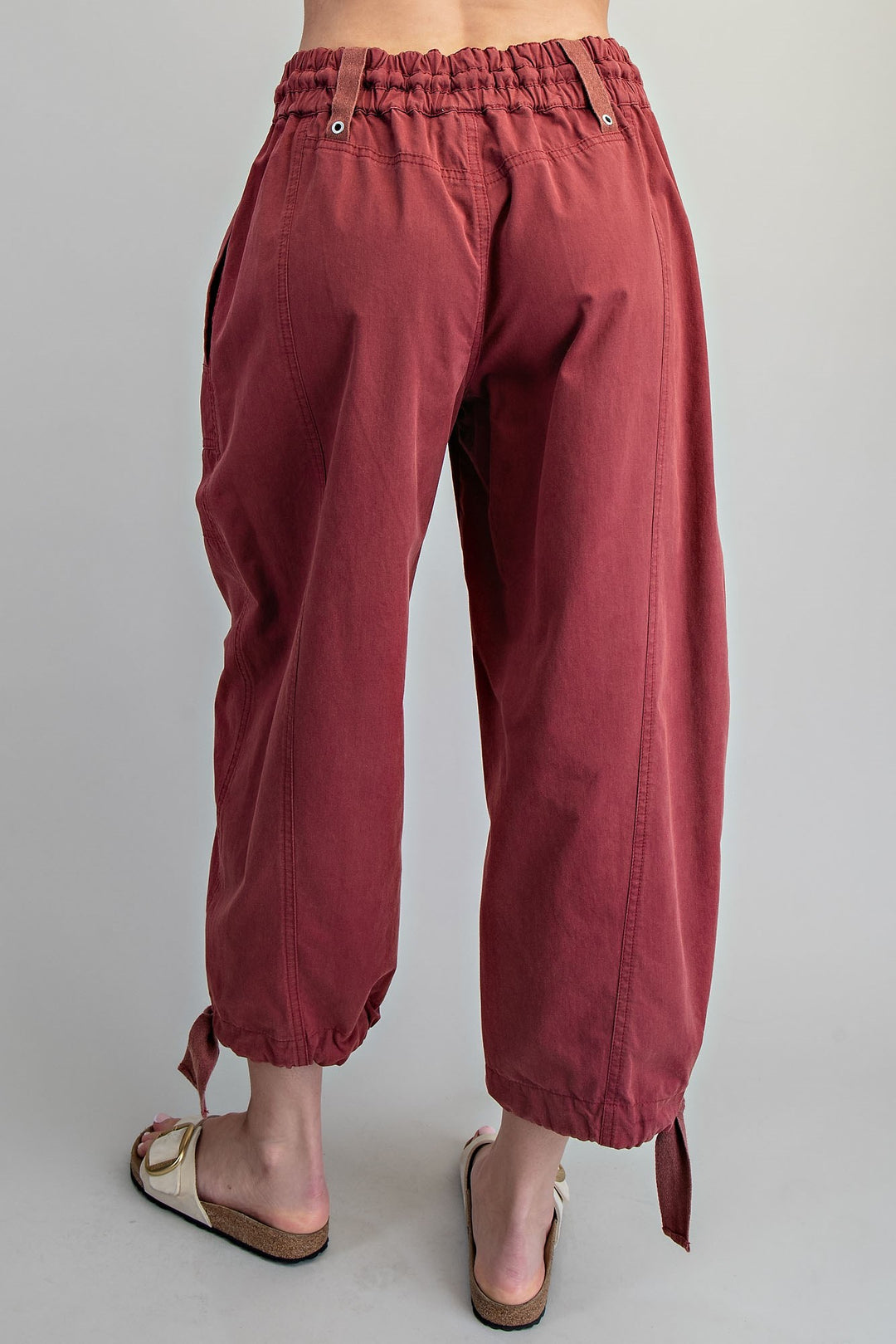 A person shown from the back wearing loose, wide-leg cropped pants in a muted burgundy or dusty rose tone, paired with beige slide sandals, against a plain light gray studio background.