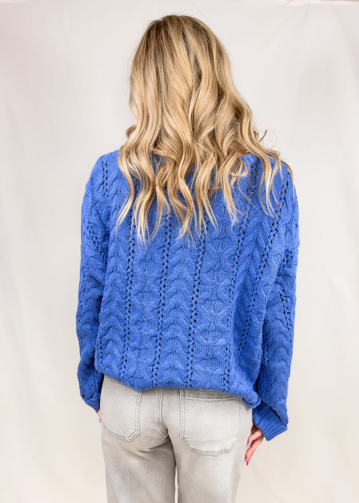 Woman shown from back wearing blue cable knit sweater on plain background