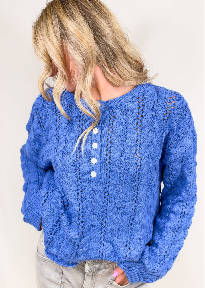 Woman shown from side view wearing blue cable knit henley pullover on plain background