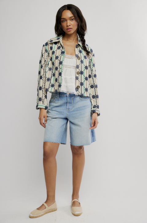 Woman wearing a patterned jacket, white top, and light blue shorts on a plain background