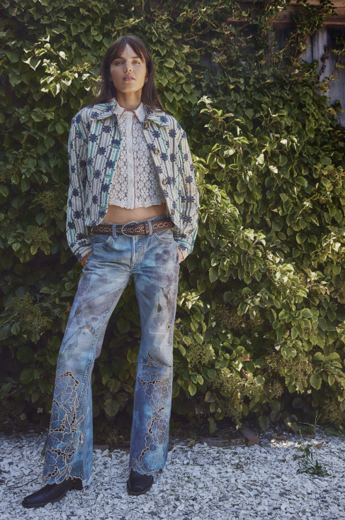 Person wearing a patterned jacket and jeans standing in front of green foliage.