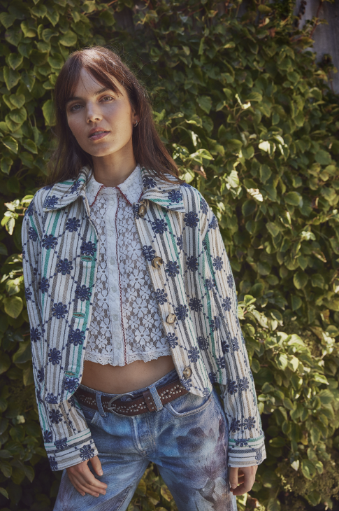 Woman wearing a patterned jacket and jeans standing against a green hedge.