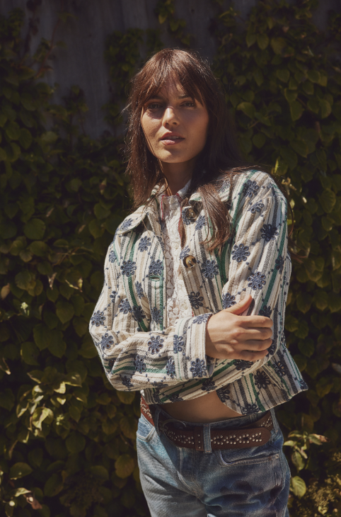 Woman wearing a patterned shirt and jeans standing against a leafy background