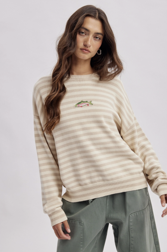 A woman wearing a cream and beige horizontally striped crewneck sweater with a small embroidered fish on the left chest, paired with olive green pants, against a light gray studio background.