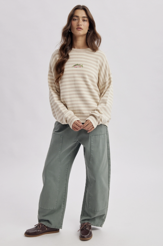 A woman modeling a cream and beige horizontally striped crewneck sweater with a small embroidered logo on the chest, paired with wide-leg sage green pants and dark brown leather shoes, against a light gray studio background.