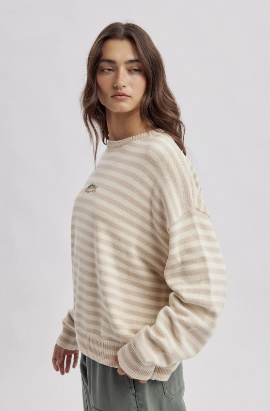 A woman modeling a cream and beige horizontally striped crewneck sweater with a small embroidered logo on the left chest, paired with olive green pants, against a light gray studio background.