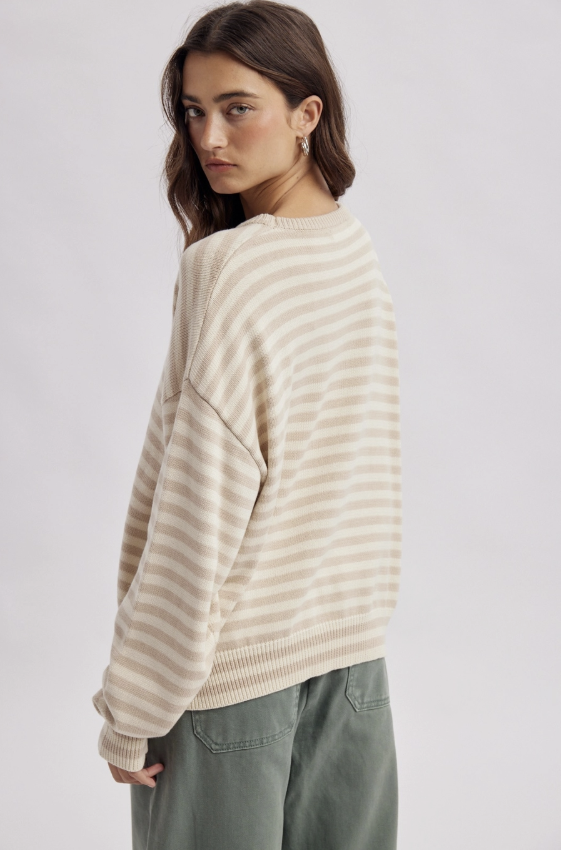 A woman wearing a cream and beige horizontally striped knit sweater, shown from a three-quarters back view against a light gray studio background.