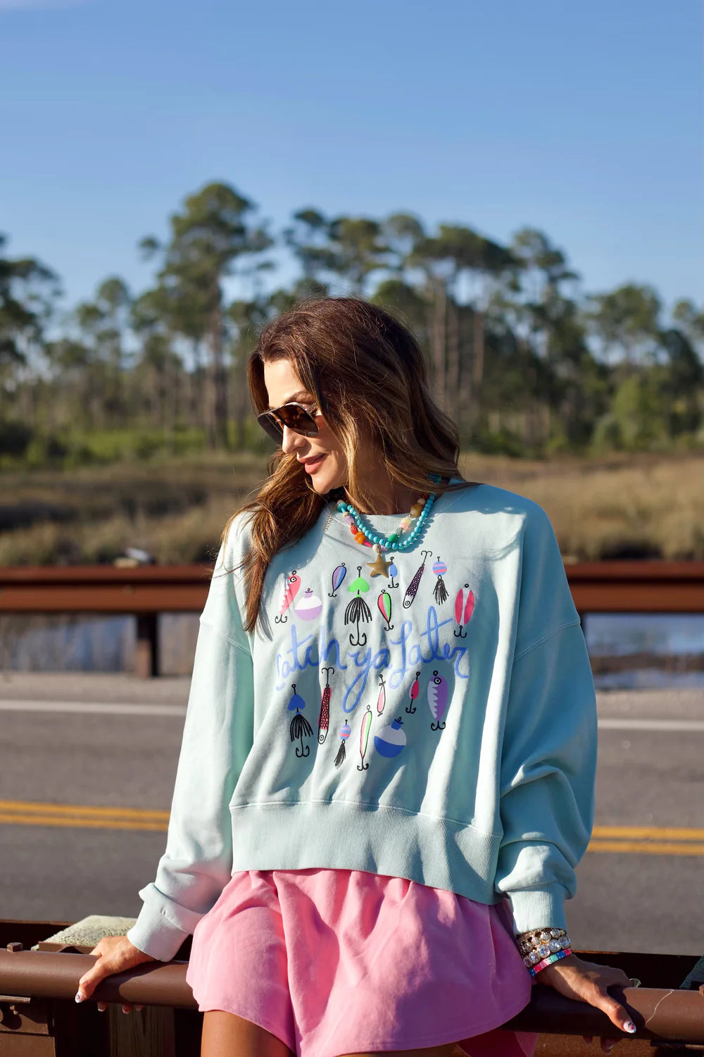 Woman wearing blue sweatshirt with fishing lure graphics and 'Catch Ya Later' writing sitting on guardrail