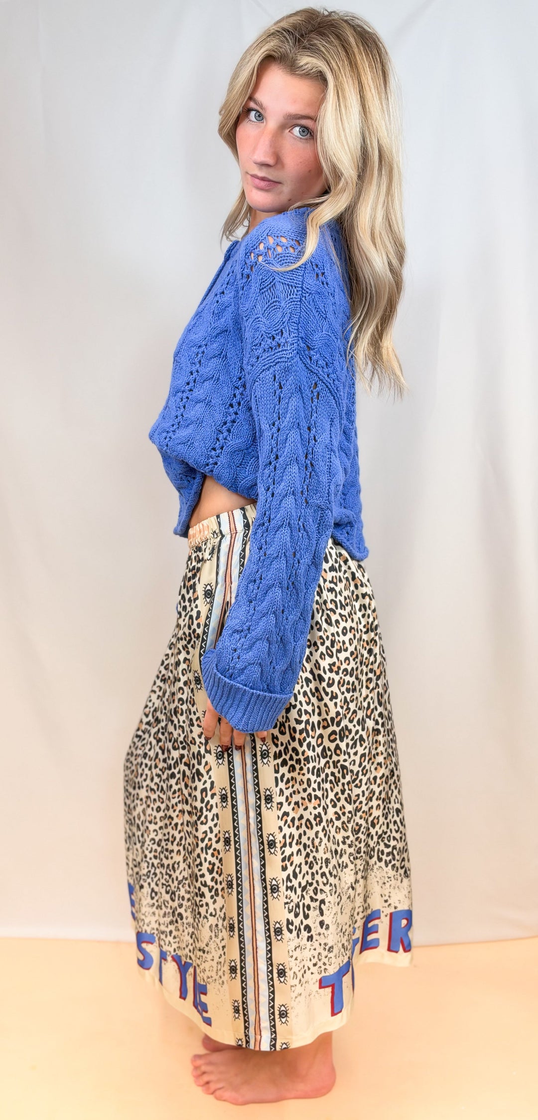 Woman shown from side view wearing blue sweater with leopard print satin skirt on plain background