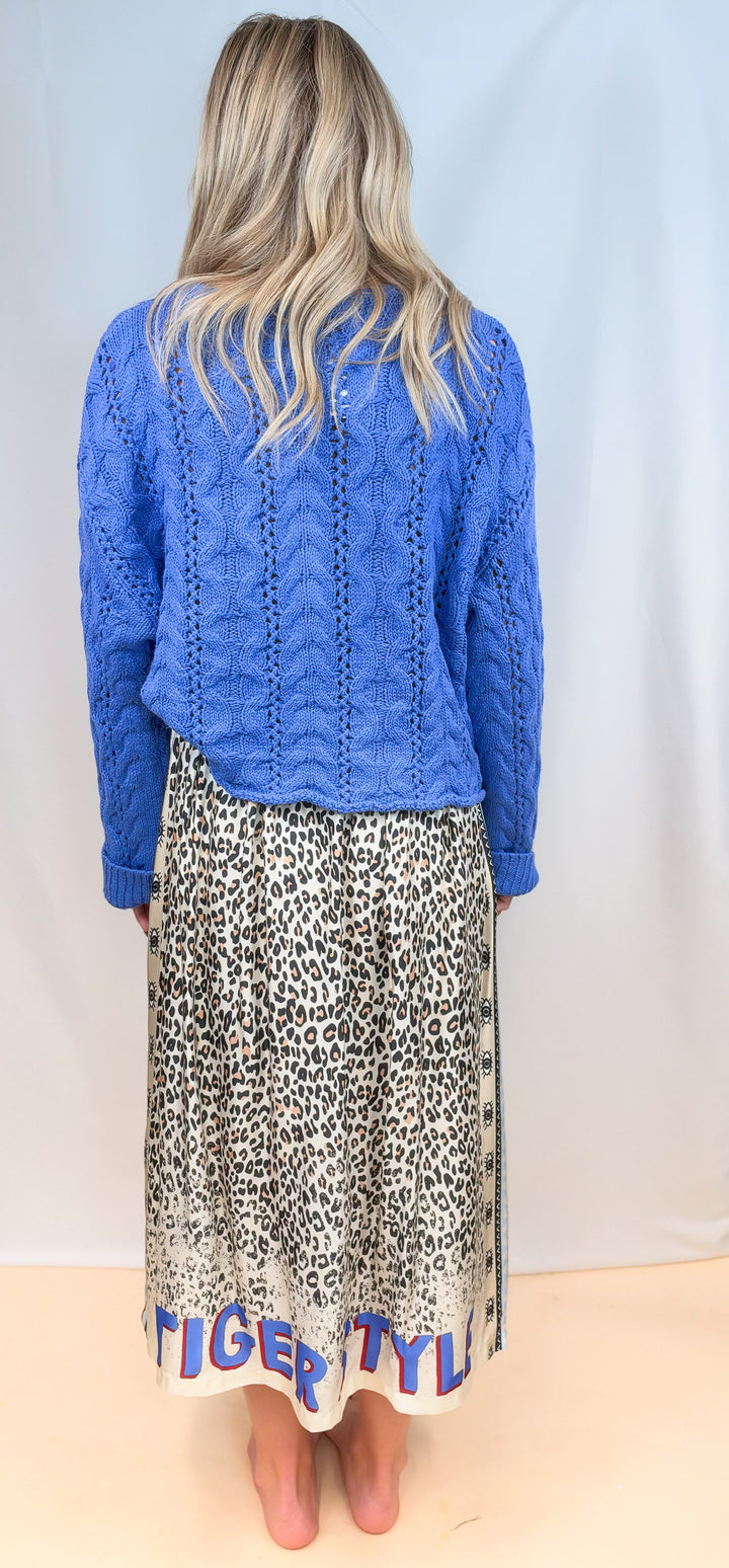 Woman shown from back view wearing blue sweater with leopard print skirt on plain background