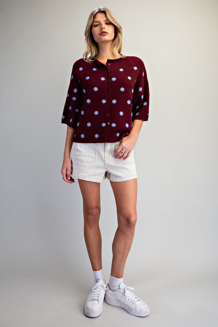 A woman modeling a burgundy velvet button-up shirt with light blue polka dots, paired with white pinstriped shorts and white sneakers, against a neutral gray studio background.
