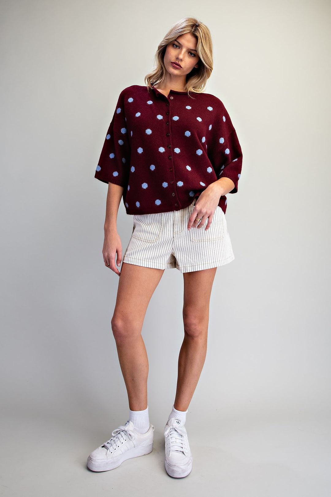 A woman modeling a burgundy short-sleeve button-up shirt with light blue polka dots, paired with white textured shorts and white sneakers, against a light gray studio background.