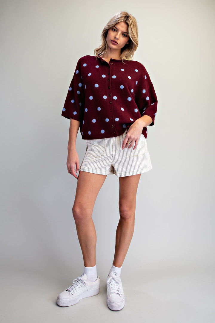 A woman modeling a burgundy short-sleeve button-up shirt with light blue polka dots, paired with white textured shorts and white sneakers, against a light gray studio background.