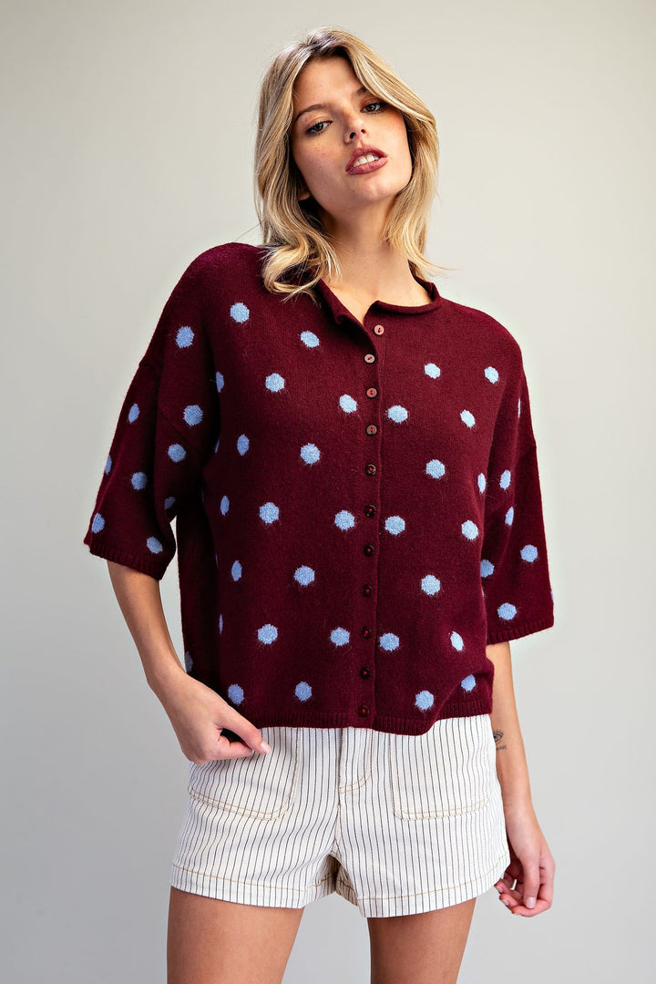A woman modeling a burgundy knit cardigan with light blue polka dots and white pinstriped shorts against a neutral studio background.
