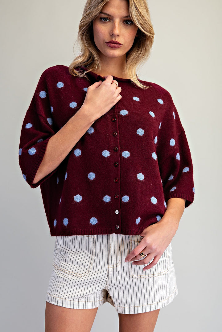 A woman modeling a burgundy knit cardigan with light blue polka dots and white pinstriped shorts against a plain light gray background.