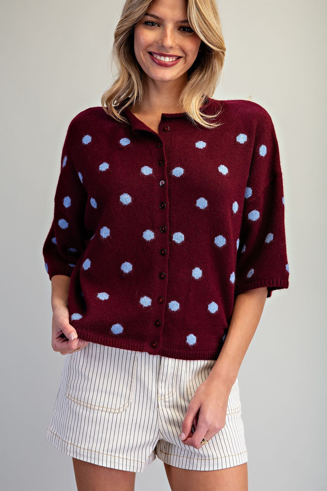 Woman wearing burgundy sweater with light blue polka dots and striped shorts on plain background