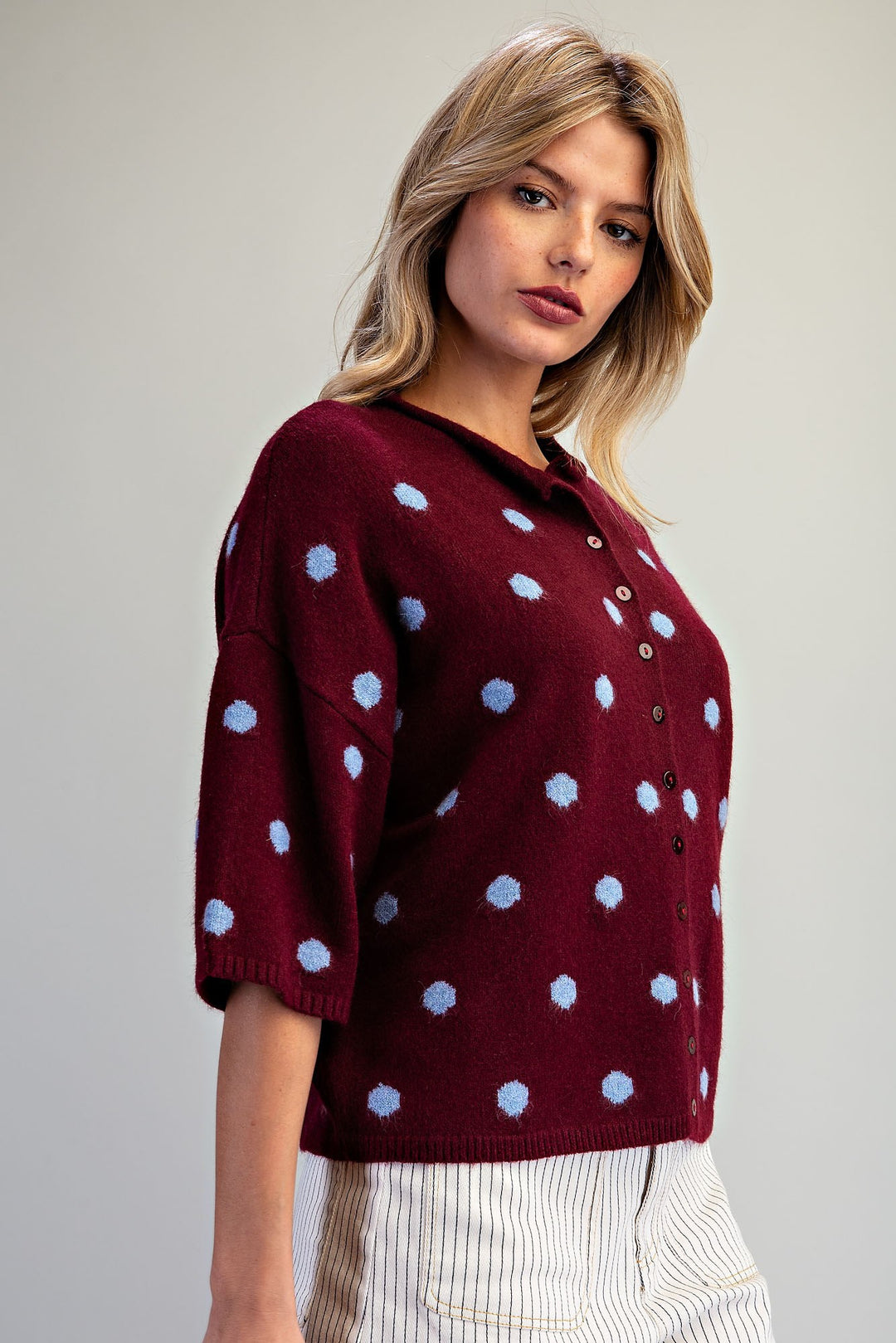 A woman wearing a burgundy knit cardigan with light blue polka dots and a buttoned collar, paired with white and navy striped trousers, against a neutral gray background.