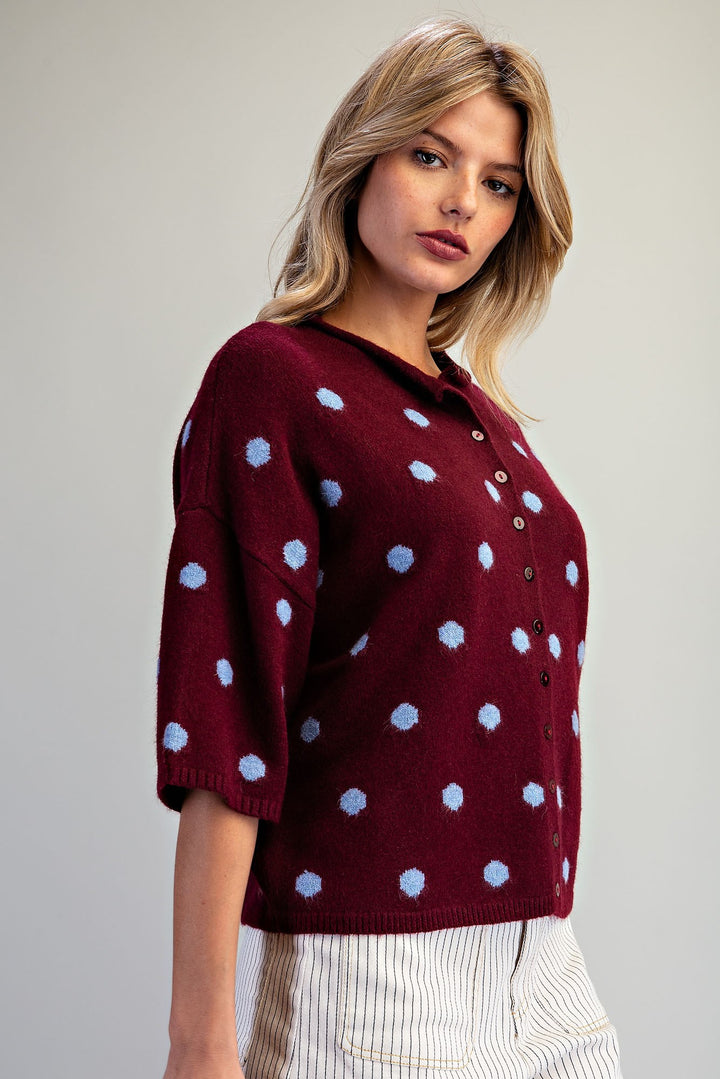 A woman wearing a burgundy knit cardigan with light blue polka dots and a buttoned collar, paired with white and navy striped trousers, against a neutral gray background.
